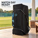 CA Pro 10000 black wheeled cricket kit bag standing beside a cricket pavilion