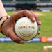 Hand holding SG Shield 30 white youth cricket ball with gold printing and dark green stitching